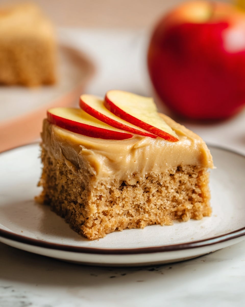 The image shows a single square piece of cake placed on a white plate with a thin dark rim. The cake has two distinct layers: the bottom layer is thick, light brown, and crumbly with a moist texture containing small bits, while the top layer is a smooth, creamy light tan frosting. On top of the frosting, there are two thin slices of red apple with yellowish skin placed at the center. The plate is on a white marbled surface with a whole red apple blurred in the background, creating a warm, cozy setting. photo taken with an iphone --ar 4:5 --v 7