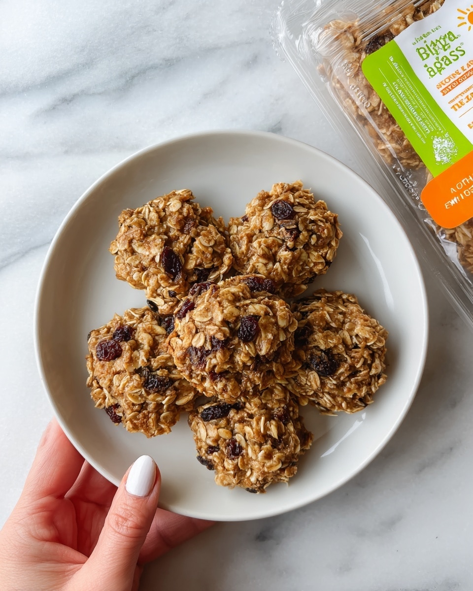 A white round plate holds five thick oatmeal bites with a rough texture, light golden brown color, and visible dark dried fruit pieces mixed throughout. The oatmeal bites are arranged close together, showing uneven, bumpy surfaces that highlight their hearty, rustic look. A woman's hand is holding one edge of the plate, with neatly manicured nails. In the background, part of a clear plastic package with a bright green and orange label is visible. The scene sits on a white marbled surface. photo taken with an iphone --ar 4:5 --v 7