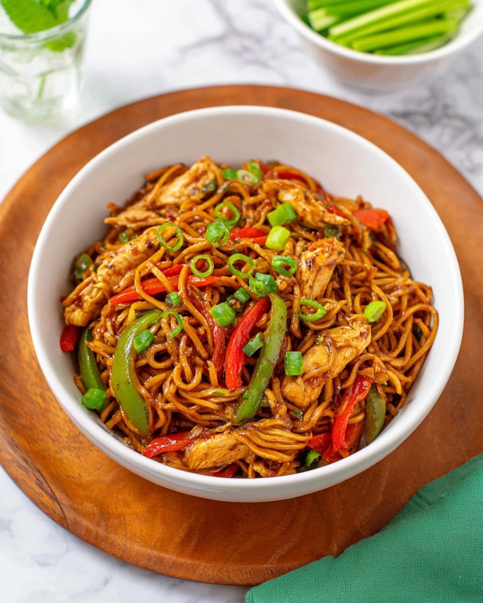 A white bowl filled with stir-fried noodles mixed with light brown cooked chicken strips, thin bright red bell pepper strips, and thin green bell pepper strips, all evenly mixed with dark brown sauce-coated noodles. The noodles and chicken are garnished with chopped green onions scattered on top. The bowl is placed on a wooden board, with a white marbled surface underneath, and a white bowl of green sliced vegetables and a glass of water with mint leaves are blurred in the background. A green cloth is visible in the lower right corner. photo taken with an iphone --ar 4:5 --v 7