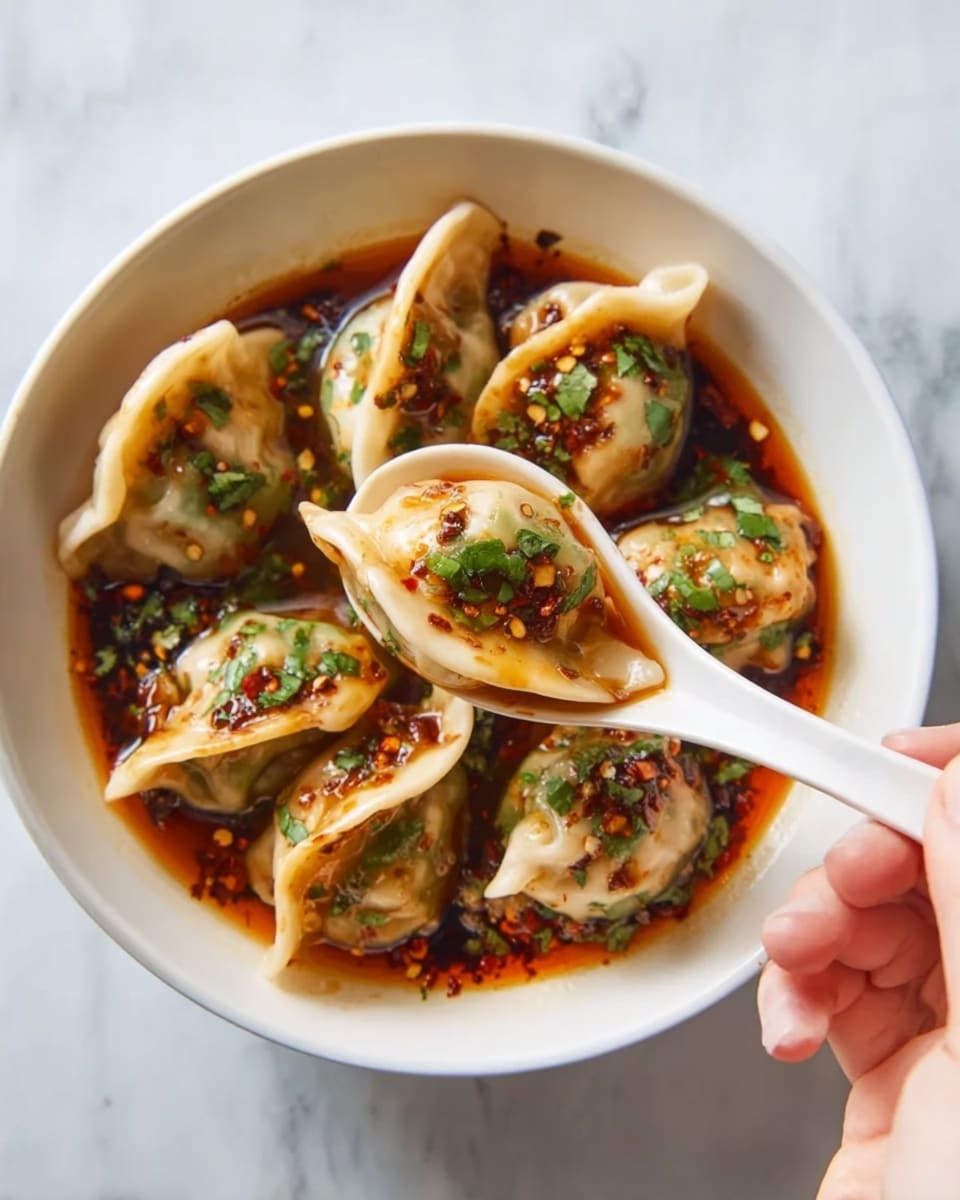 A white bowl holds seven dumplings in a dark reddish-brown sauce with visible chili flakes and chopped green herbs. Each dumpling has a light beige, soft wrapper showing a slightly wrinkled texture with green herbs on top and sauce glistening on them. A woman's hand holds a white ceramic spoon lifting one dumpling, highlighting its plump shape and the shiny sauce covering it. The background is a white marbled surface. photo taken with an iphone --ar 4:5 --v 7