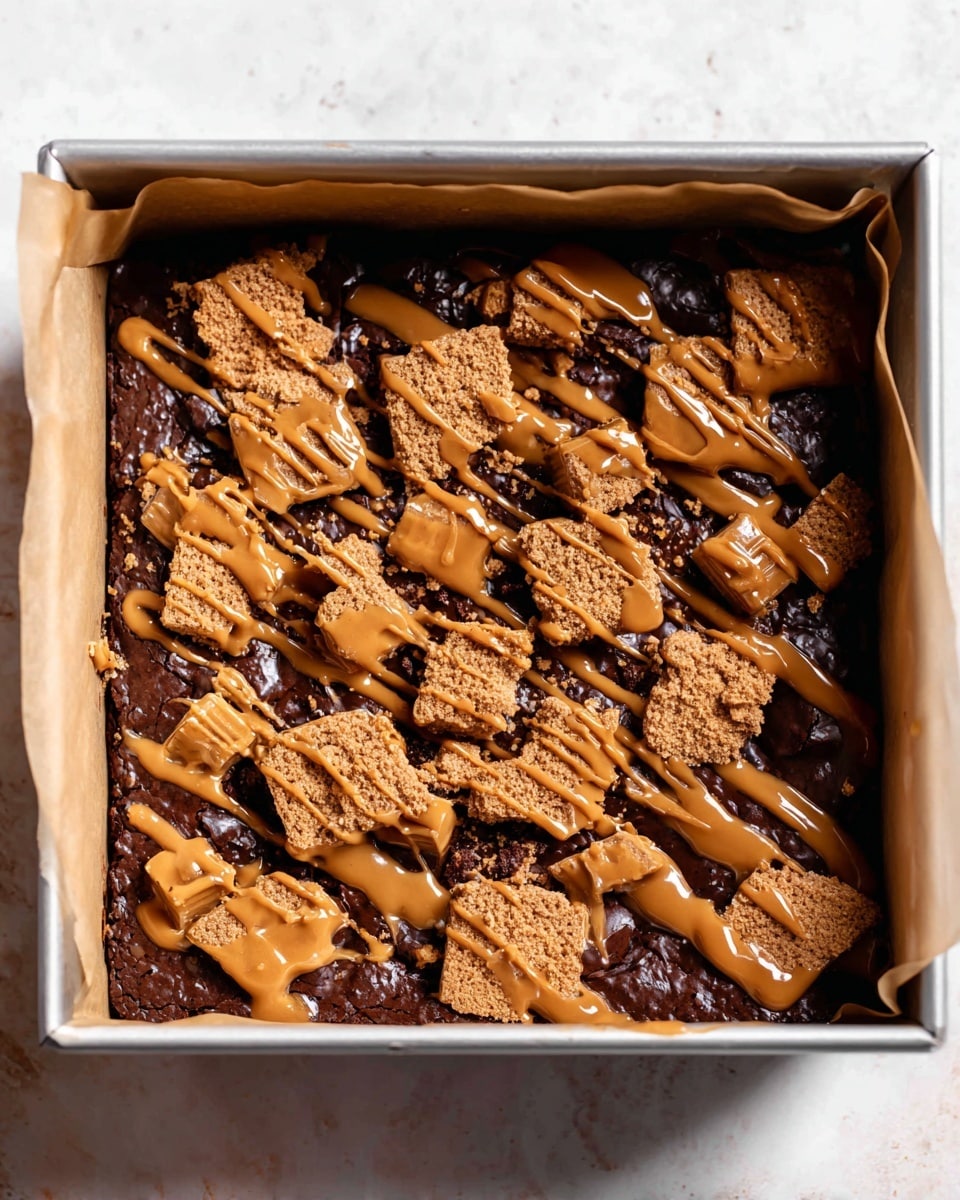 A square baking pan lined with parchment paper holds a rich, dark chocolate brownie layer as the base. On top, there are large pieces of light brown biscuit scattered unevenly across the surface, creating a rough texture with small crumbs in between. A caramel-colored sauce is drizzled generously over the biscuits in thin, irregular lines. The pan rests on a white marbled surface. photo taken with an iphone --ar 4:5 --v 7