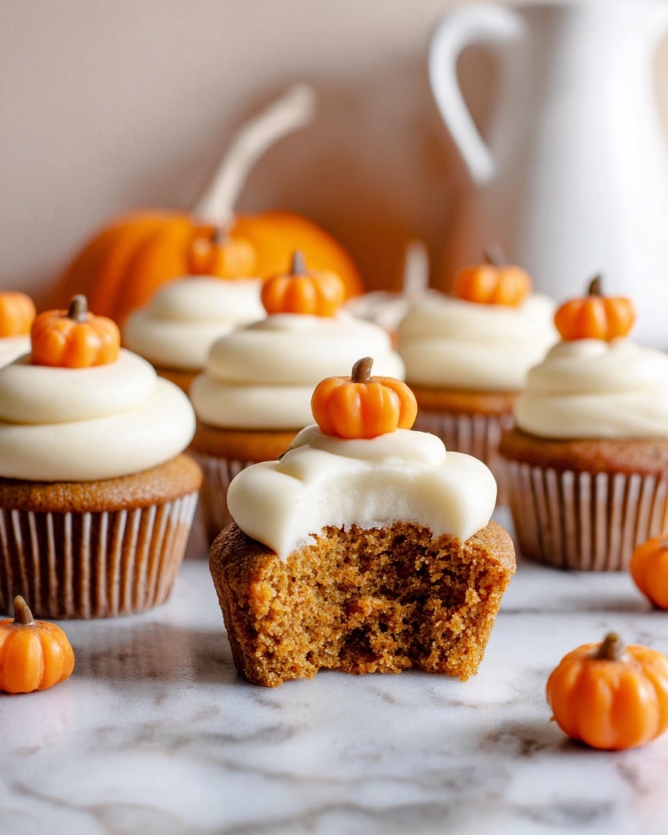 This image shows several small cupcakes with two clear layers. The bottom layer is a soft, golden brown cake with a slightly crumbly texture. On top, there is a thick, creamy white frosting swirled smoothly around each cupcake. Each frosting swirl is decorated with a small, bright orange pumpkin-shaped candy placed on the top center. One cupcake in the middle has a bite taken from it, showing the inside of the cake and frosting. The cupcakes sit on a white marbled surface with extra pumpkin candies scattered around. Photo taken with an iphone --ar 4:5 --v 7