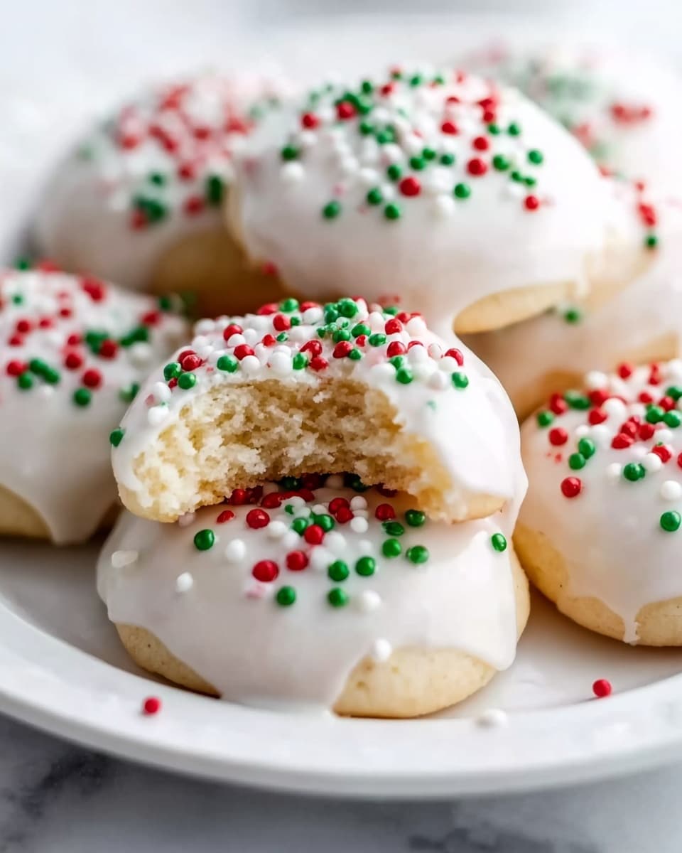 A white plate holds several small round cookies covered in smooth white icing that drips slightly over the edges. The cookies are topped with small round red, green, and white sprinkles, adding a festive look. One cookie is in front with a bite taken out, showing a soft, light, and slightly crumbly inside texture. The background is a white marbled surface, enhancing the clean and bright feel of the scene. photo taken with an iphone --ar 4:5 --v 7