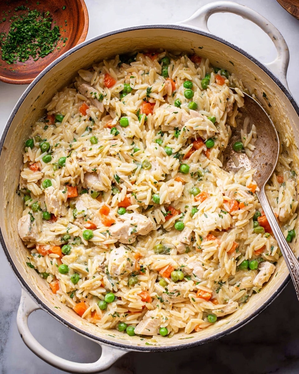 Large white enamel pot filled with creamy orzo pasta cooked with tender chunks of chicken, bright green peas, diced carrots, and aromatic herbs, the mixture glistening under natural light, a rustic silver spoon resting inside, whole dish captured from a slightly angled overhead view on a white marble countertop, styled professionally like a food magazine hero shot, photo taken with an iphone --ar 4:5 --v 7