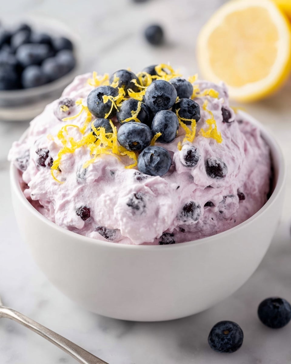 Large white bowl brimming with creamy blueberry lemon fluff salad, a light and airy mixture studded with whole blueberries and flecks of lemon zest, topped with a generous pile of fresh blueberries and additional lemon zest for garnish, whole dish presented on a white marble countertop with natural lighting, professional 3/4 angle hero food shot, photo taken with an iphone --ar 4:5 --v 7