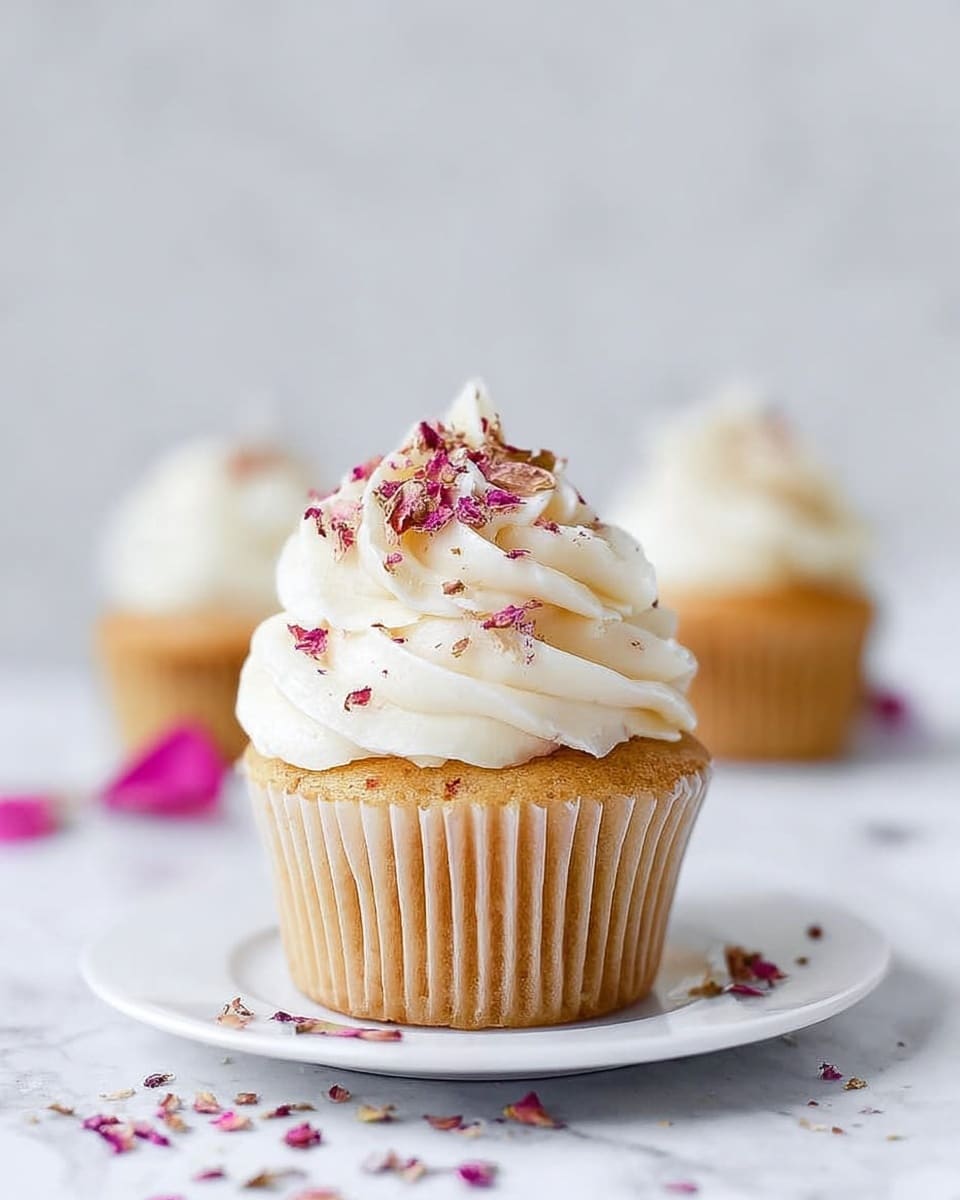 A pristine white rectangular serving platter presenting a full batch of twelve delicately baked vanilla cupcakes, each topped generously with swirls of smooth white frosting and sprinkled with elegant dried rose petals. The cupcakes are uniformly arranged in neat rows, showcasing their light golden bases and fluffy texture. The background features a clean white marble countertop with soft natural lighting enhancing the gentle colors and textures, styled like a high-end food magazine hero shot. photo taken with an iphone --ar 4:5 --v 7