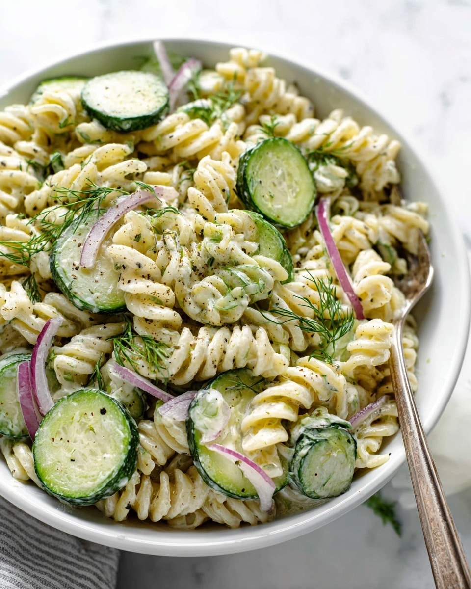 Large white oval serving dish filled with creamy cucumber pasta salad, featuring al dente rotini pasta tossed with thinly sliced crisp cucumbers and delicate red onion rings, all coated in a smooth, herb-speckled dressing, garnished generously with fresh dill sprigs and freshly ground black pepper, presented on a white marble countertop under natural light, professional overhead food magazine style photo taken with an iphone --ar 4:5 --v 7
